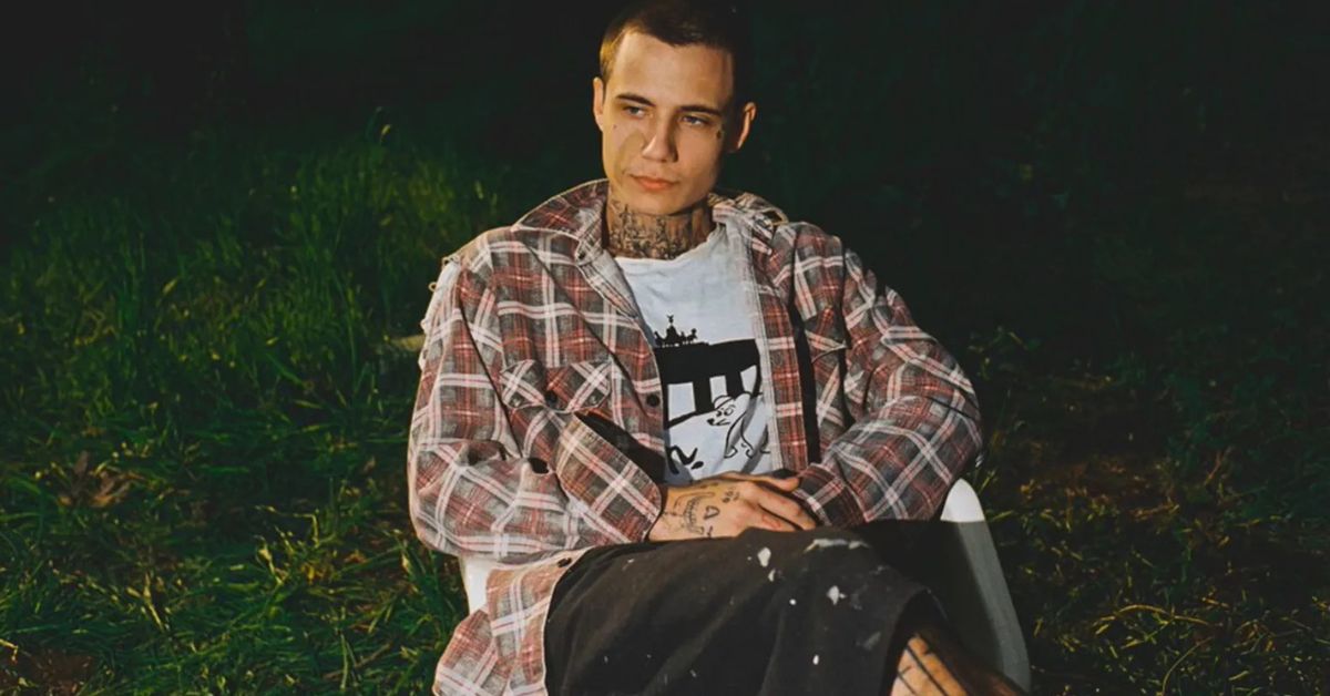 Jxdn (Jaden Hossler) photographed sitting on a white plastic lawn chair with a grassy area behind him, he is wearing casual clothing with a flannel shirt, white t-shirt with a cartoon dog and black mid-length shorts with paint splashes upon them. 