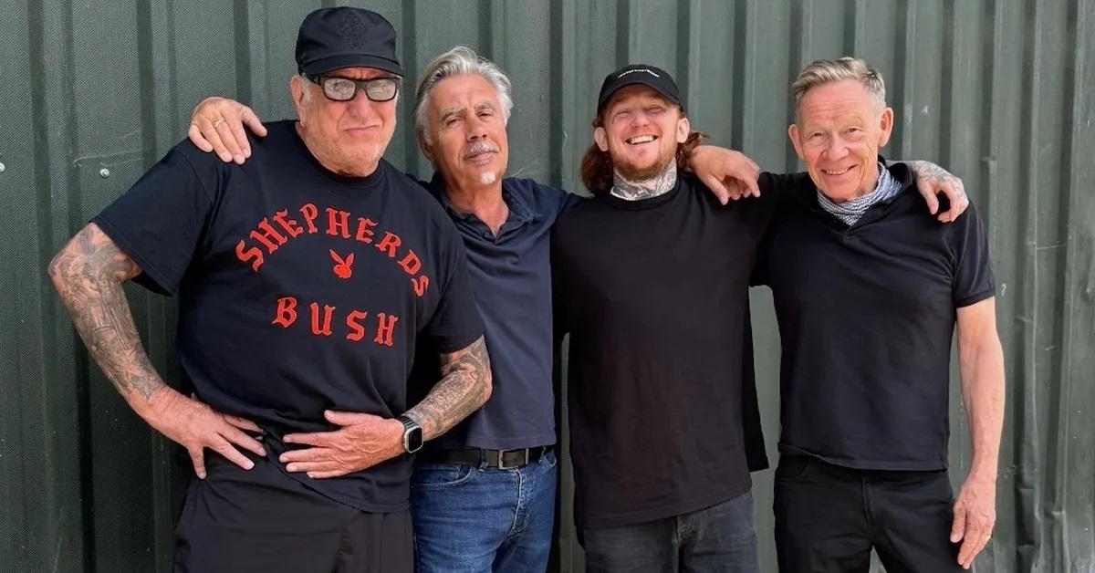 Sex Pistols members, photographed with Frank Carter, standing against a green metal fence, they are all wearing dark t-shirts and jeans