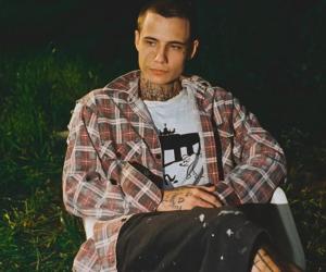 Jxdn (Jaden Hossler) photographed sitting on a white plastic lawn chair with a grassy area behind him, he is wearing casual clothing with a flannel shirt, white t-shirt with a cartoon dog and black mid-length shorts with paint splashes upon them. 