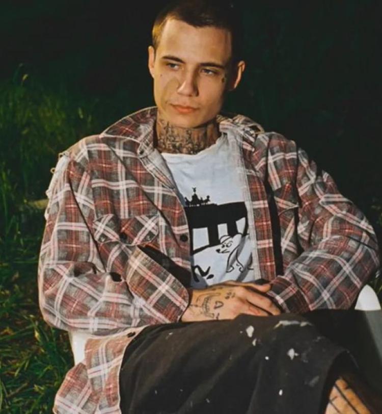 Jxdn (Jaden Hossler) photographed sitting on a white plastic lawn chair with a grassy area behind him, he is wearing casual clothing with a flannel shirt, white t-shirt with a cartoon dog and black mid-length shorts with paint splashes upon them. 